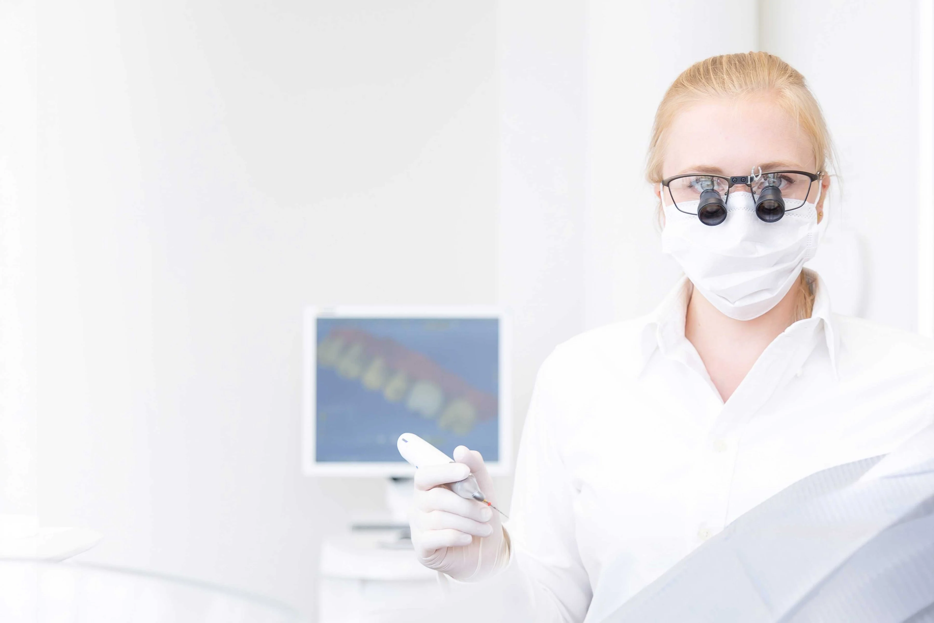 Behandlerin mit Lupenbrille und Intraoralscanner vor Monitor mit 3D-Scan